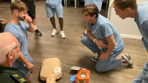 Tom Williams/BBC Men in blue kits surround a dummy used for CPR training as an emergency worker looks on.